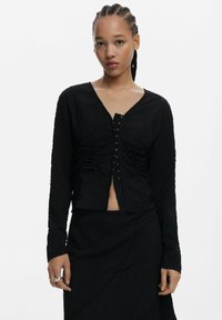 Top nero strutturato con maniche lunghe, scollatura a V e chiusura frontale con bottoni decorativi in metallo. Abbinato a una gonna nera coordinata.