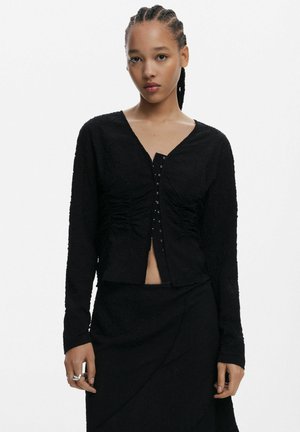 Top nero strutturato con maniche lunghe, scollatura a V e chiusura frontale con bottoni decorativi in metallo. Abbinato a una gonna nera coordinata.