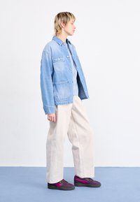 Veste en jean bleu clair avec deux poches avant, associée à un haut bleu clair et un pantalon ample crème. Chaussures violet foncé avec des accents roses.