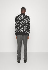 Man som står vänd mot en vägg iförd en svart tröja med vit textdesign "Just Cavalli", grå byxor och svarta skor.