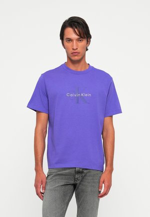 Man wearing purple Calvin Klein logo t-shirt and grey jeans standing against plain light background.