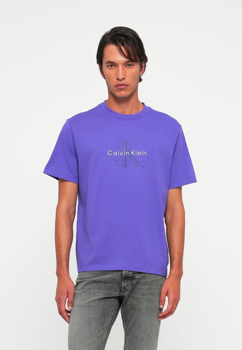 Muž v purpurovom tričku s logom Calvin Klein a šedých džínsoch stojí proti jednému svetlému pozadiu.