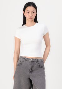 Hvit kortermet crop-top laget av et glatt stoff, kombinert med høytlivs grå denimbukser med frontlommer og knappelukking.