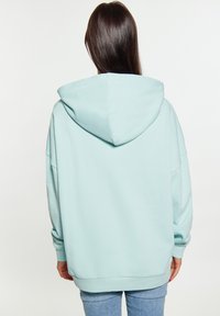 Svetlomodrá oversized mikina s kapucňou na šnurovanie. Má rebrované manžety a lem, a mäkkú textúru. Pohľad zozadu ukazuje voľný strih.