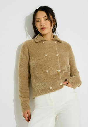 Mujer con un suéter doble botonadura beige y peludo con cuello y pantalones blancos, posando contra un fondo claro.