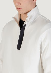 Suéter blanco con un cuello alto que presenta un acento de cremallera negra y un inserto negro contrastante en la abertura del cuello. Textura suave.
