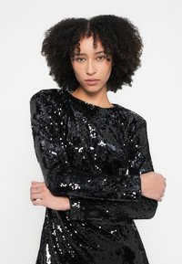 Melns spīguļojošs vakarkleita ar garām piedurknēm, šauru piegriezumu, struktūru virsmu, apaļu kakla izgriezumu un vieglu drapējumu vidukļa zonā.