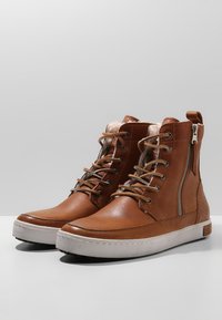 Botas de tobillo de cuero marrón con lados texturizados, cremallera metálica y parte delantera con cordones. Cuenta con un interior acolchado y suela de goma blanca.