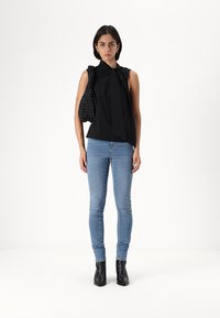 Svart ärmlös topp med en rosettdetalj, tillsammans med ljusblå smala jeans och svarta ankelstövlar. Modellen håller en texturerad svart väska.