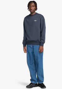 Felpa blu marina con polsini e orlo a coste, caratterizzata da un logo bianco sul petto, abbinata a jeans oversize azzurri e scarpe nere.