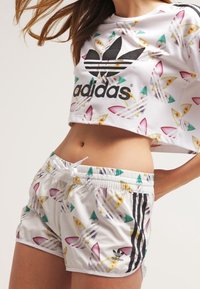 Bílý crop top s barevným vzorem surfboardů a černým logem Adidas. Odpovídající šortky mají stejný vzor, černé pruhy a elastický pas se šňůrkou.