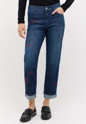 Person iført mørke opsmøgede jeans med farverige broderede designs, sort top og sorte loafers, vist fra skuldre til fødder mod en ensfarvet baggrund.