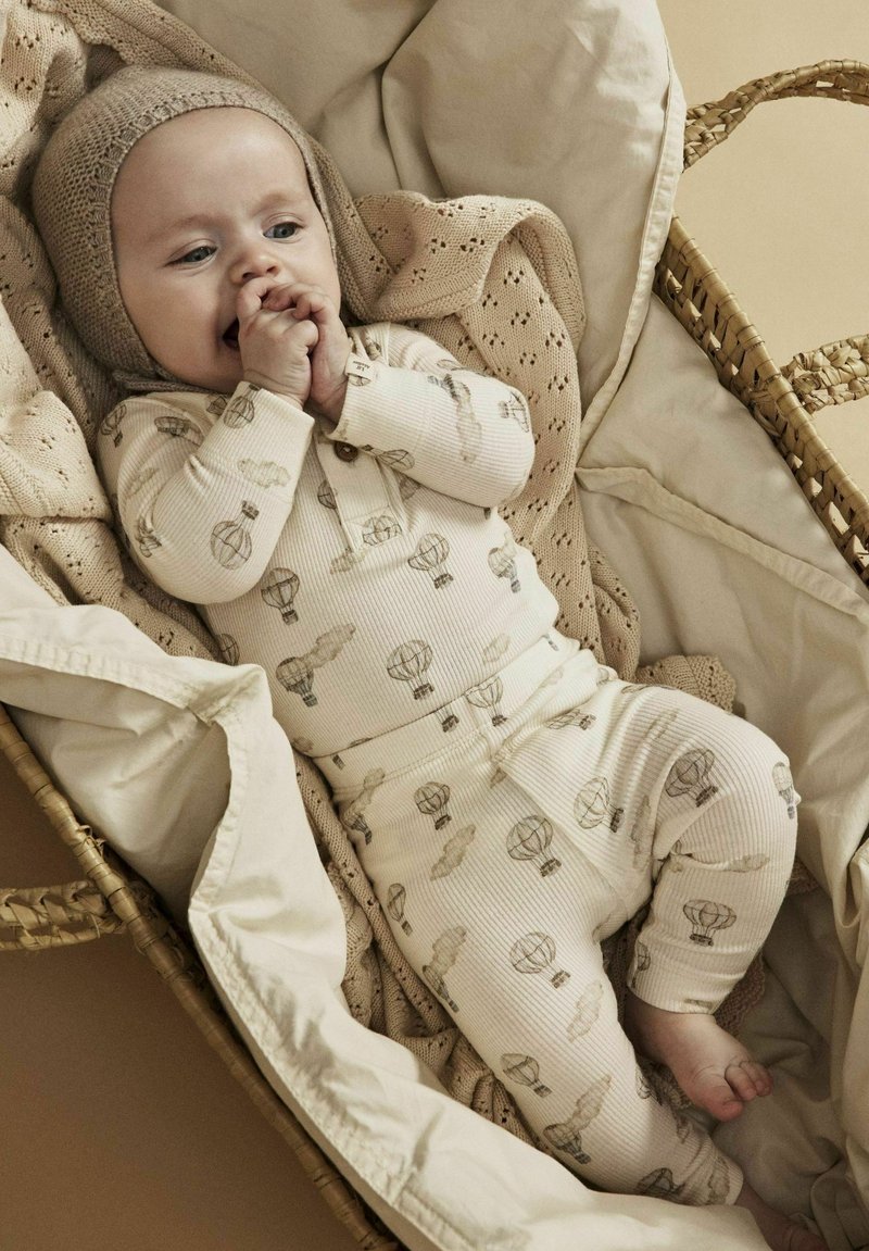 Pigiama in cotone leggero color crema con una texture a costine, caratterizzato da una stampa di palloncini giocosa. Il bambino è in un cesto intrecciato foderato con una coperta lavorata a maglia.