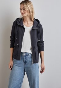 Felpa navy con zip e finitura testurizzata, cappuccio con coulisse regolabile, due tasche frontali e fodera liscia. Abbinata a jeans azzurri.