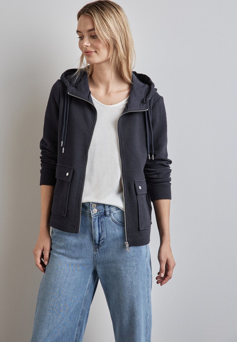 Felpa navy con zip e finitura testurizzata, cappuccio con coulisse regolabile, due tasche frontali e fodera liscia. Abbinata a jeans azzurri.