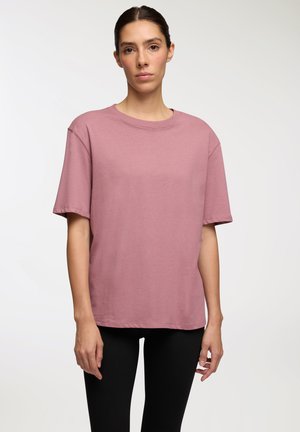 T-shirt di cotone rosa con vestibilità rilassata, scollo rotondo e maniche corte, abbinato a leggings neri. Texture liscia, dettagli minimi.