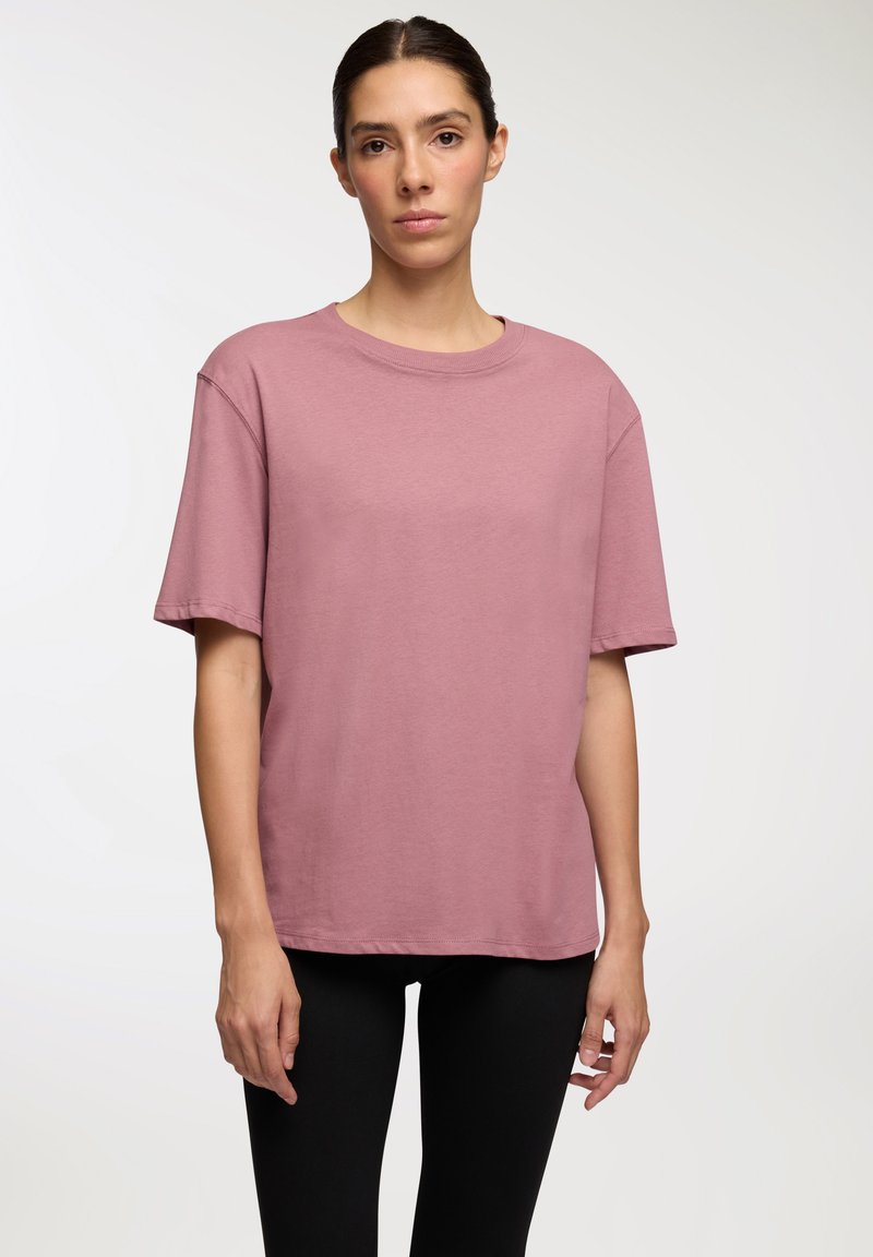 Różowa bawełniana koszulka z luźnym krojem, okrągłym dekoltem i krótkimi rękawami, w połączeniu z czarnymi legginsami. Gładka faktura, minimalne detale.