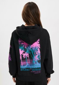 Črna kapuca z barvnim grafičnim motivom palm na hrbtni strani, z napisom "HOLIDAY DIVISION". Mehak material in sproščen kroj.