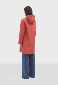 Cappotto rosso fino al ginocchio con cappuccio, caratterizzato da una linea dritta e fessure laterali. Indossato sopra pantaloni blu a righe, vista da dietro.