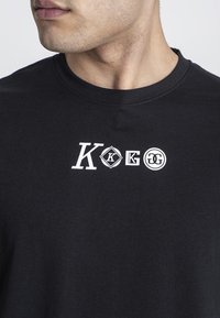 Černé bavlněné tričko s kulatým výstřihem, na přední straně bílé grafické potisky písmen a symbolů. Jednoduchý a minimalistický design.