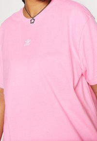 Person som bär en ljusrosa Adidas-t-shirt med en liten vit logotyp på bröstet och ett svart halsband med en blommig berlock.