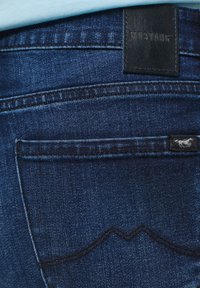 Detailní záběr na zadní kapsu modrých džín s černou značkovou nálepkou Mustang a embosovanou logovou nášivkou nad kapsou.