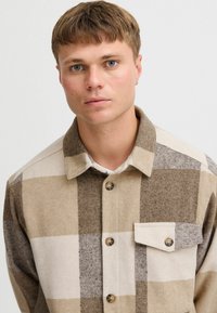Camicia a quadri marrone e beige, realizzata in tessuto testurizzato, con chiusura a bottoni, colletto e tasca sul petto con dettaglio a bottone.