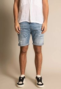 Shorts de denim, azul claro, con detalles desgastados y dobladillos enrollados. Combinados con una camisa de lino blanca y zapatillas en blanco y negro.