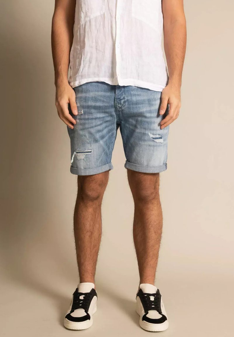 Shorts de denim, azul claro, con detalles desgastados y dobladillos enrollados. Combinados con una camisa de lino blanca y zapatillas en blanco y negro.