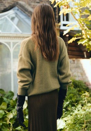 Suéter de punto verde oliva con hombros caídos, combinado con una larga falda marrón con pliegues. Guantes de cuero negros completan el conjunto.