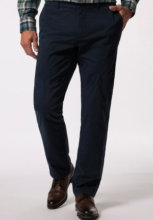 Homme portant un pantalon bleu marine, des chaussures en cuir marron et une chemise à carreaux verte, une main dans la poche, debout sur un fond uni.