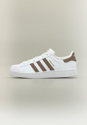 Baskets Adidas Superstar blanches avec des bandes, des lacets et une languette de talon marron sur fond beige neutre, vue de côté.