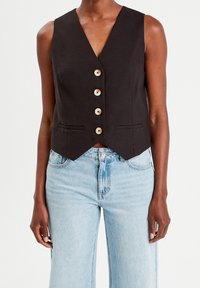 Gilet noir sans manches avec un col en V, doté de cinq boutons en bois et de deux poches avant. Le tissu a une texture lisse, conçu pour une coupe ajustée.