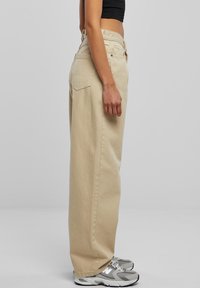 Béžové kalhoty s širokými nohavicemi vyrobené z bavlny, s vysokým pasem, bočními kapsami a volným střihem, doplněné šedými teniskami.