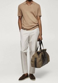 Personne portant un polo court beige, un pantalon beige clair, des mocassins marron, tenant un grand sac de sport olive et marron foncé par ses poignées.