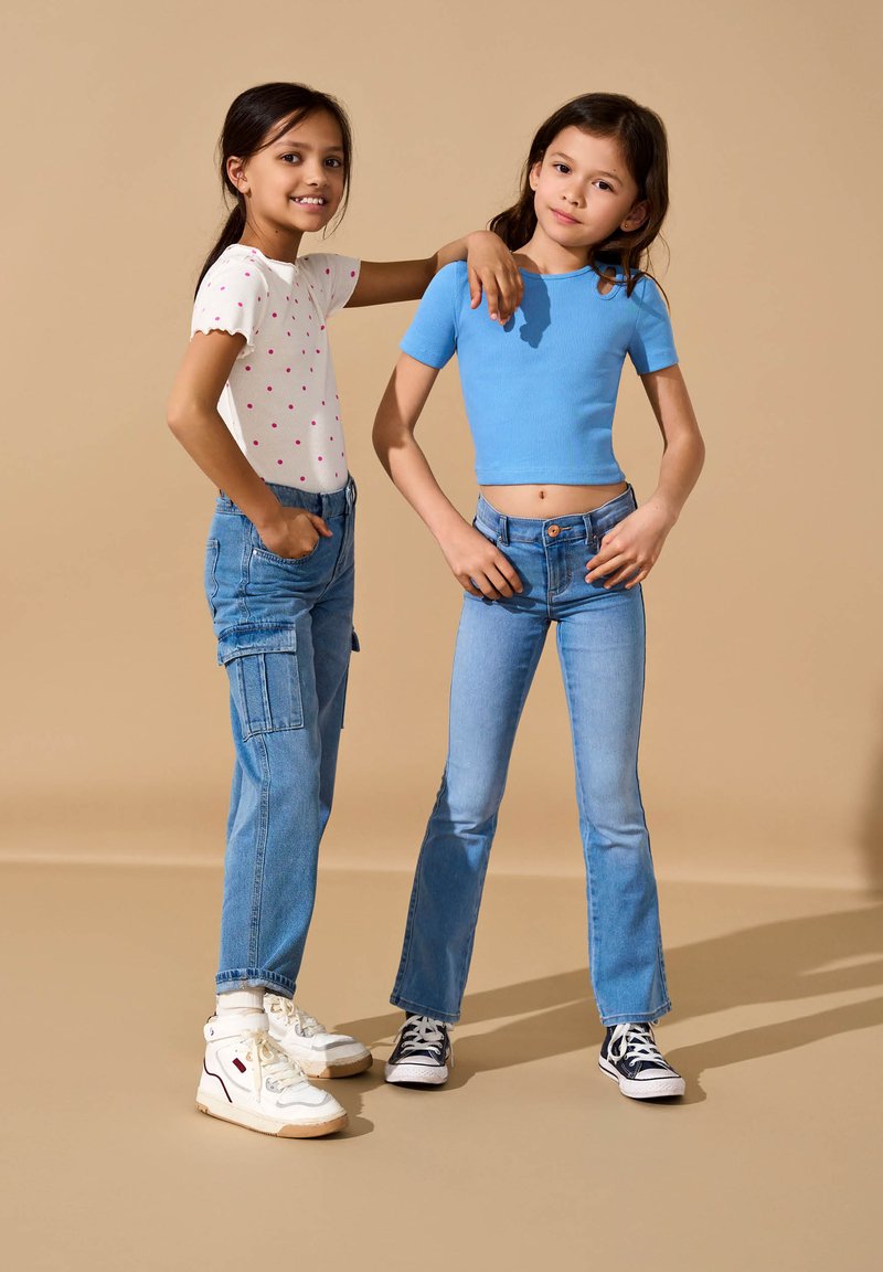 Duas crianças estão juntas contra um fundo bege. Uma usa uma camisa branca com bolinhas rosas e jeans cargo; a outra usa um top azul curto e jeans flare azul claro.