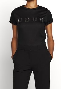 Camiseta negra de manga corta con logotipo "HUGO" en relieve y reflectante. Combinada con pantalones negros ajustados, que muestran una textura suave.