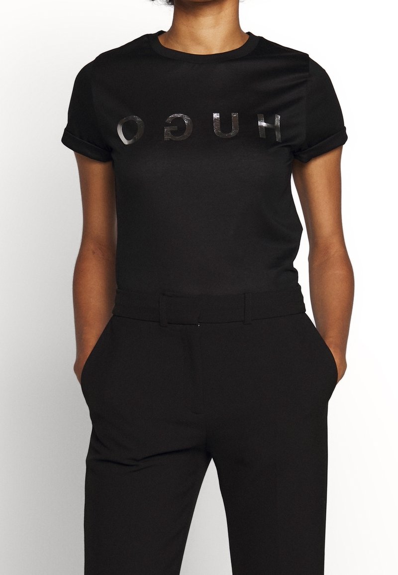Camiseta negra de manga corta con logotipo "HUGO" en relieve y reflectante. Combinada con pantalones negros ajustados, que muestran una textura suave.