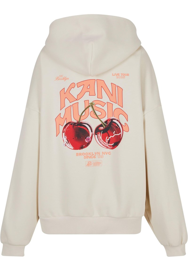 Kreemist kapuutsiga kampsun, mille graafikal on kaks kirsit, ja "KANI MUSIC" tugeva oranži fondiga, sisaldab kangaroo taskut ja soonikkoes varrukaotsasid.