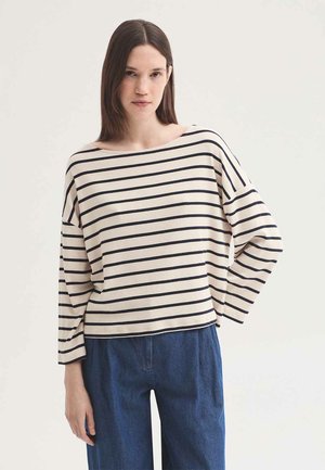 Young woman with straight brown hair wearing a white long-sleeve top with black horizontal stripes and blue high-waist jeans.