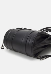 Sac à main cylindrique en cuir noir posé sur le côté avec une bandoulière détachable et des ferrures argentées sur fond blanc.