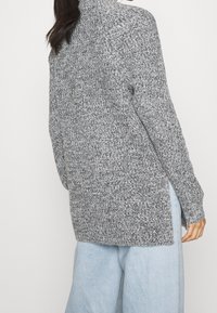 Grå stickad tröja med hög krage, texturerat mönster och sidofrakturer, tillsammans med ljusblå vida byxor.