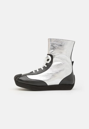 Vyšší tenisky vyrobené z černé kůže a lesklého stříbrného materiálu. Mají bílé tkaničky a texturovanou černou gumovou podrážku.