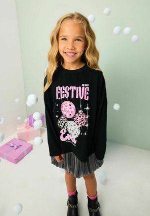 Lachend meisje met lang blond haar, dragend een zwarte feestelijke trui, grijze rok, roze sokken en zwarte laarzen in een versierde kamer met cadeaus.