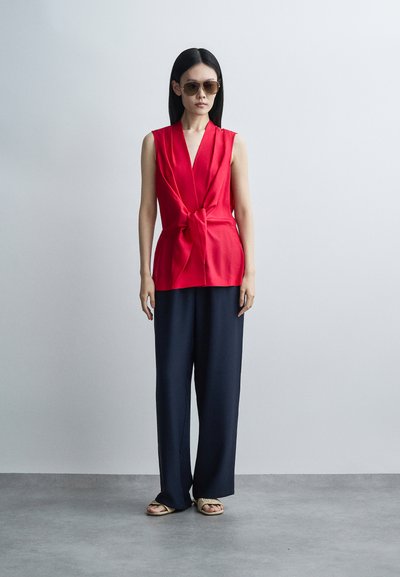 Femme aux longs cheveux noirs portant des lunettes de soleil marron, un haut rouge sans manches avec un nœud devant, un pantalon large noir et des sandales beige, debout sur un sol gris.