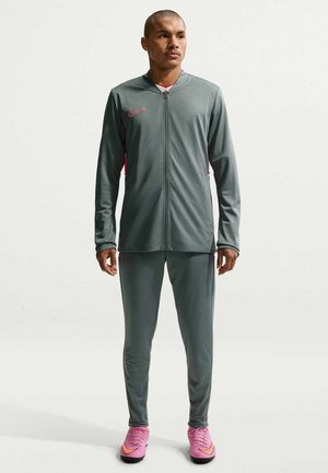 Mannequin masculin portant une veste de sport grise à fermeture éclair intégrale et un pantalon assorti avec des chaussures Nike roses, debout devant un fond blanc, faisant face à l’avant.