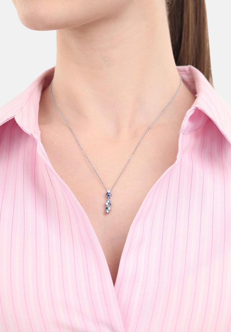 Silvrig kedja med en vertikal hänge av växelvis blå och vita ädelstenar som bärs av en person i en rosa randig skjorta.