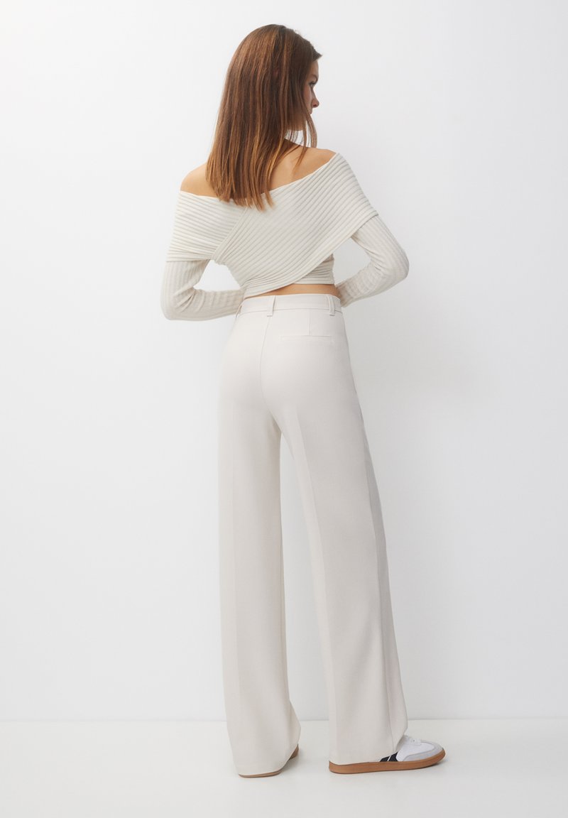 Pantaloni ampi di colore beige chiaro con una texture liscia, caratterizzati da una vestibilità sartoriale e una tasca posteriore. Abbinati a un top a coste off-the-shoulder.