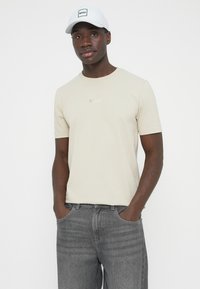 Svetlo bež kratka majica z okroglim izrezom, z nežno logotipom "BOSS" v ujemajoči se barvi nad prsmi. Kombinirana s temno sivimi kavbojkami.