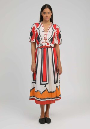 Femme debout portant une robe mi-longue aux manches courtes bouffantes avec des motifs géométriques rouges, noirs, orange et blancs. Des ballerines noires complètent la tenue.